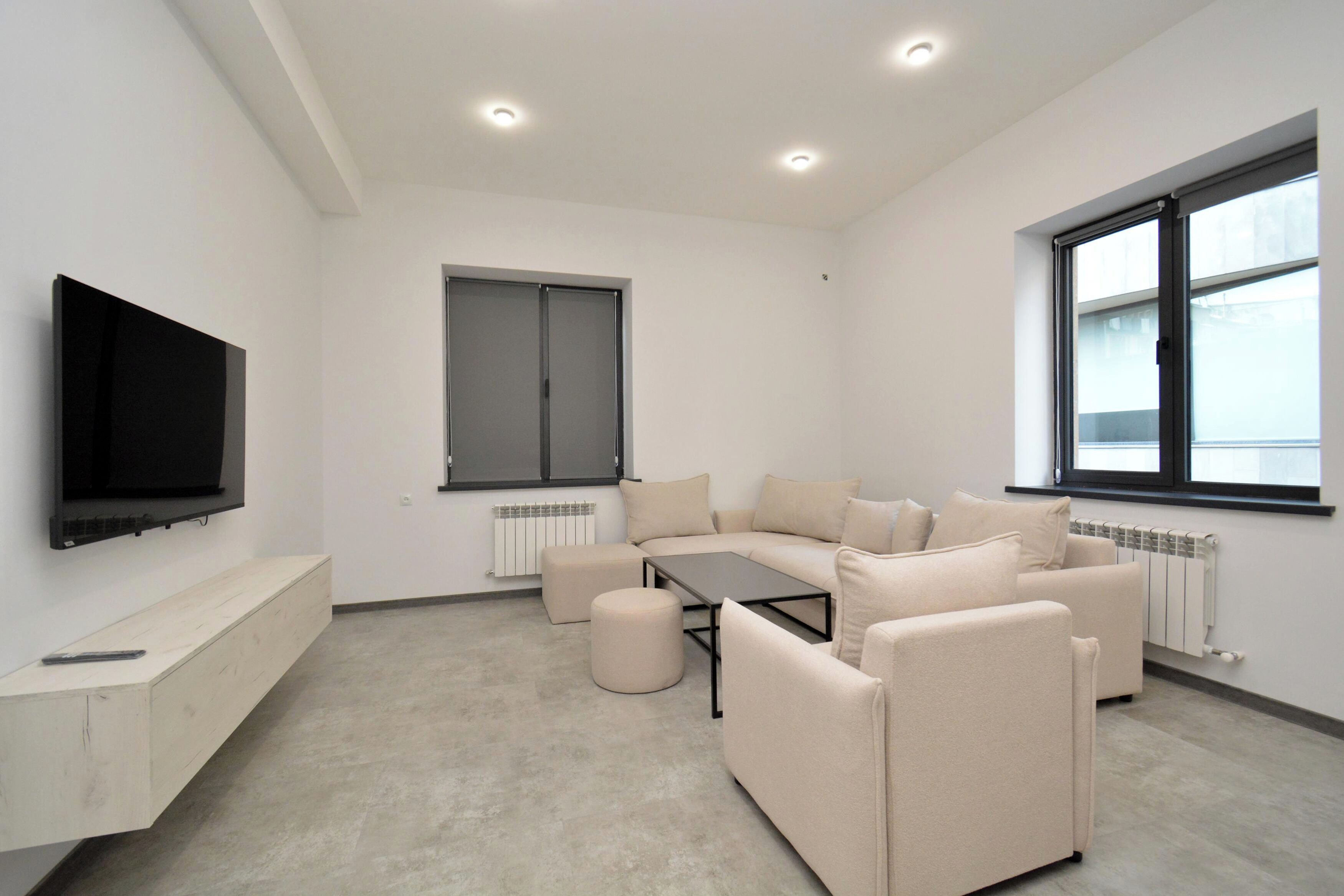 Ein modernes Wohnzimmer mit einer Sofagarnitur, einem Couchtisch, einem an der Wand befestigten Flachbildfernseher und einem Fenster, das natürliches Licht hereinlässt.