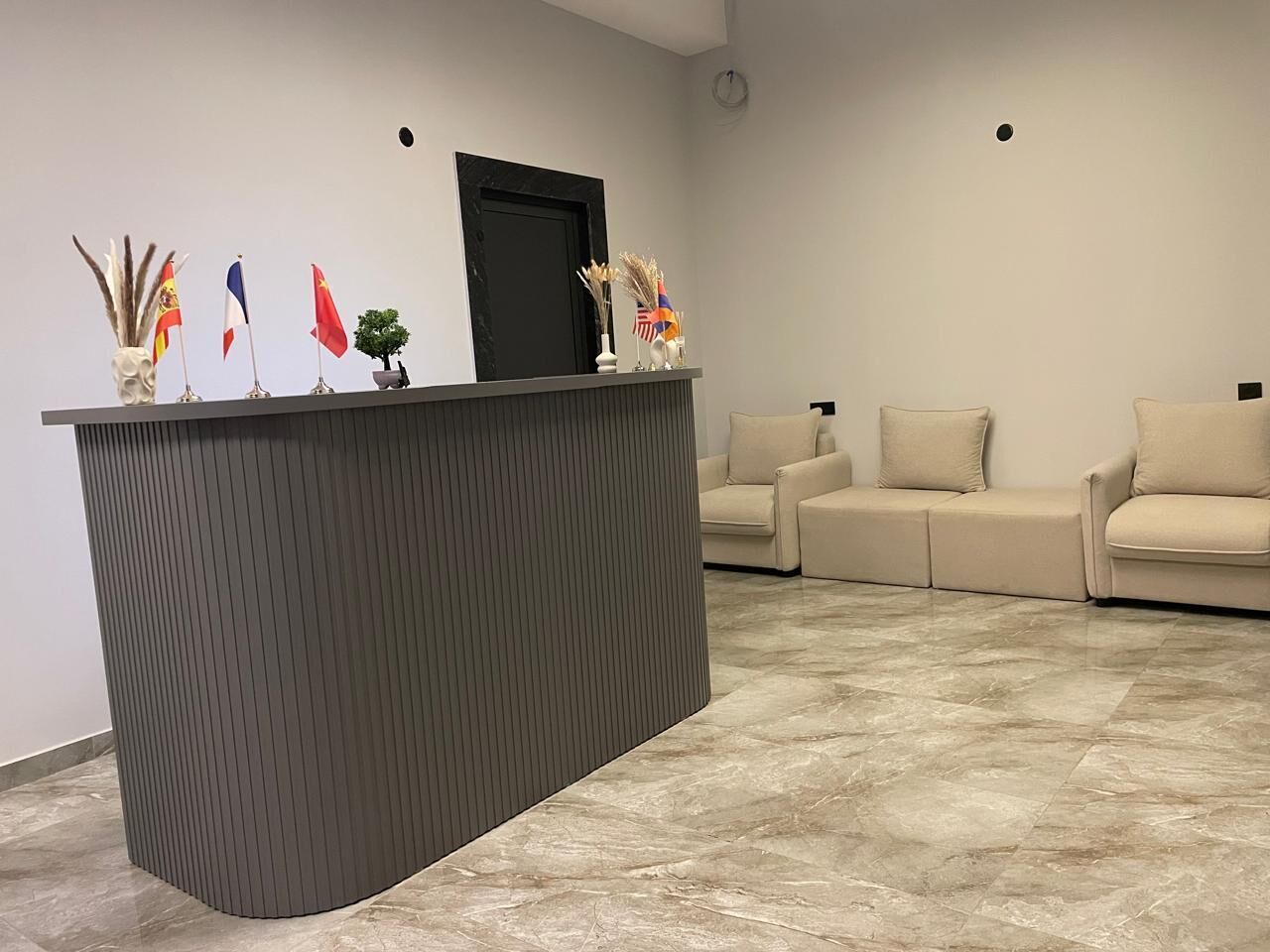 Un comptoir d’accueil orné de drapeaux, une porte noire et un canapé beige dans un hall d’entrée moderne.