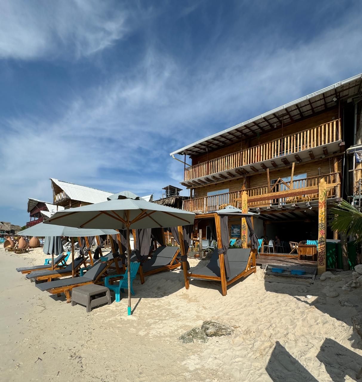 A beachside building with multiple levels, outdoor seating with umbrellas, and a sandy beach area.
