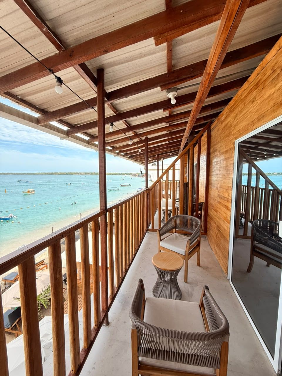 Un balcón techado con barandillas de madera, mobiliario de mimbre y vistas a la playa.