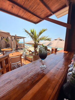 A wooden bar with a glass of dark-colored drink, outdoor seating, and a view of the beach. - w cabañas beach (Cartagena)
