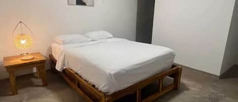 Un dormitorio con cama de madera, ropa de cama blanca, una mesita de noche con lámpara y un cuadro colgado en la pared.