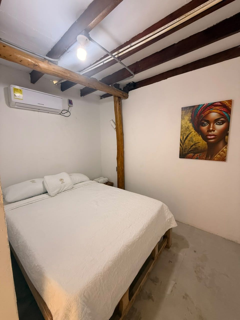 A bedroom with a bed, an air conditioning unit, a wooden beam ceiling, and a painting on the wall.