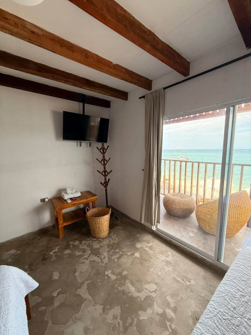 A room with a television, a wooden bench, a wicker basket, and a view of the beach through a sliding door.
