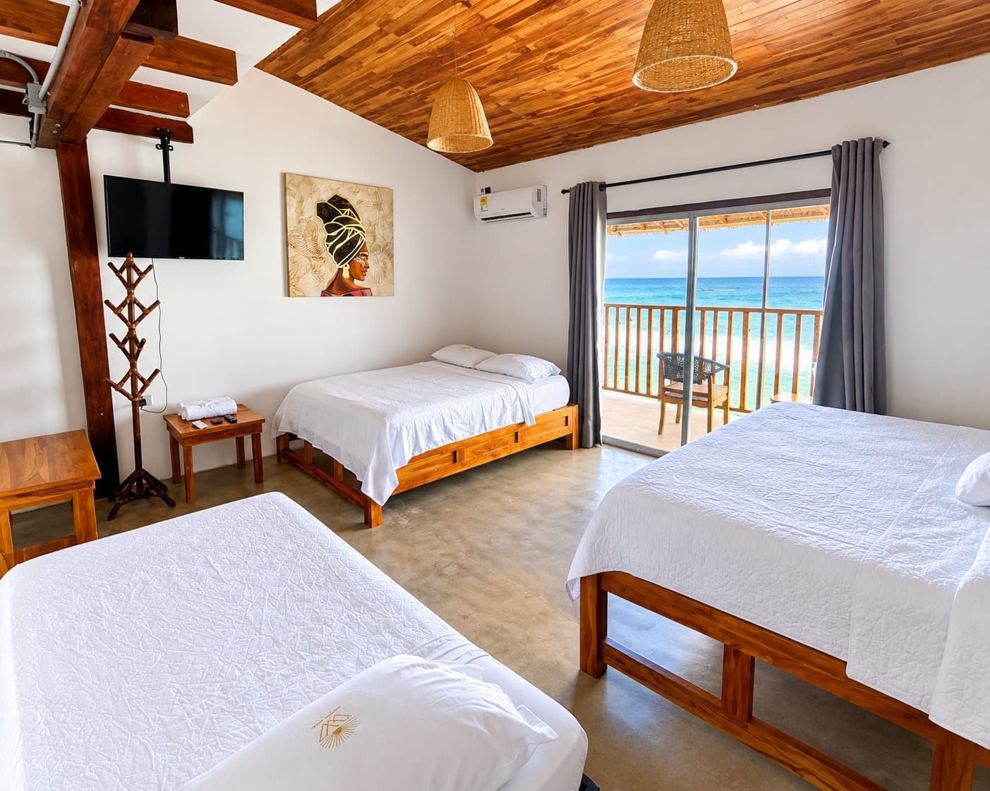 A hotel room with two beds, a TV, a painting, and a balcony with a view of the sea.