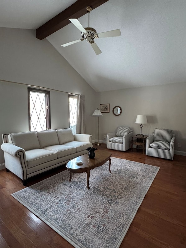 A living room with a sofa, two armchairs, a coffee table, a patterned rug, a ceiling fan, and a window with curtains.