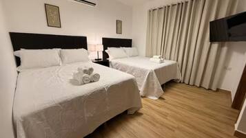 A hotel room with two beds, a flat-screen TV, a window with curtains, and an air conditioning unit.