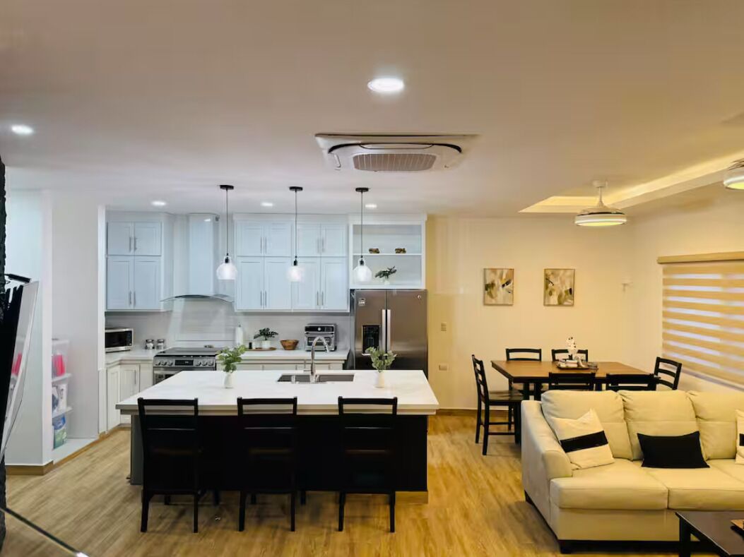 A modern kitchen with white cabinets, a central island with chairs, a dining area with a table and chairs, and a living room with a sofa and a coffee table.
