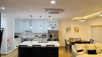 A modern kitchen with white cabinets, a central island with chairs, a dining area with a table and chairs, and a living room with a sofa and a coffee table.