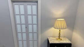 Ein Zimmer mit Holzboden, einer weißen Tür mit Milchglasfenstern, einem kleinen schwarzen Nachttisch mit Schubladen und einer Lampe mit gestreifter Lampenschirm.