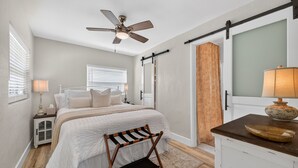 Quarto com cama, mesa de cabeceira, escrivaninha, cadeira, ventoinha no teto e porta de correr.