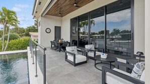 Eine Terrasse im Freien mit Sitzgelegenheiten, einem Pool und großen Fenstern mit Blick auf Palmen und einen klaren Himmel.