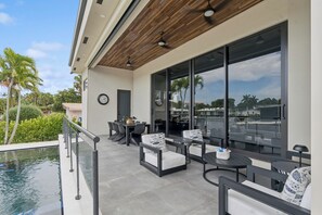 Eine Terrasse im Freien mit Sitzgelegenheiten, einem Pool und großen Fenstern mit Blick auf Palmen und einen klaren Himmel.