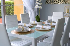Una mesa de comedor dispuesta con sillas blancas, platos y servilletas, una orquídea en maceta y un adorno en la pared.