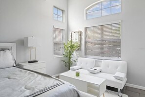 Un dormitorio moderno con cama, mesita de noche, lámpara, sofá blanco y ventanas amplias con persianas.