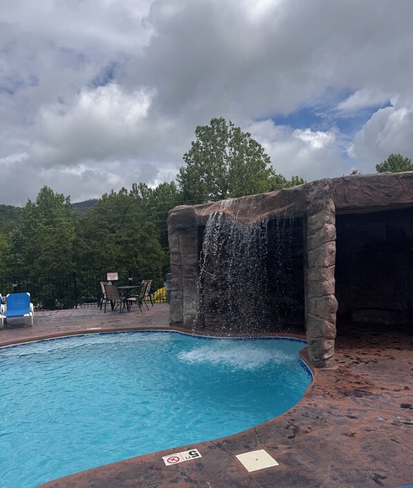 A swimming pool with a waterfall feature, surrounded by stone walls and a patio area with chairs. - Cozy 3 Bd Cabin 20 Mins to Dollywood, Fireplace, Private Hot Tub, Golf Arcade (Gatlinburg)