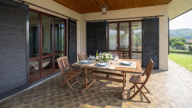 An outdoor dining area with a table set for four, surrounded by wooden chairs, on a patio with a tiled floor.