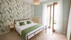 A bedroom with a bed, a bedside table, a radiator, a window with curtains, and a view of the outdoors.