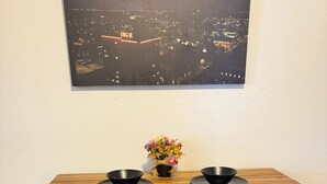 Een eettafel met twee zwarte krukken, een vaas met bloemen en een stadsgezichtschilderij aan de muur.