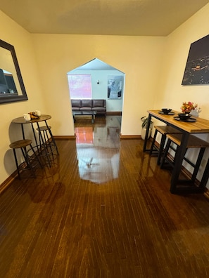 Una habitación con piso de madera, un sofá, una mesa de comedor con sillas, una mesita redonda con taburetes y un cuadro colgado en la pared.