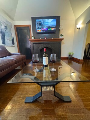 Una sala de estar con una mesa de centro de vidrio, una chimenea, un televisor montado encima y un sofá de cuero marrón.