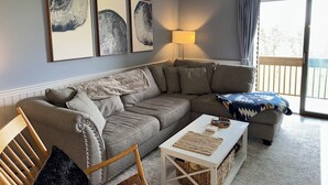 Una sala de estar con un sofá gris, una mesa de centro blanca y una silla de madera. Hay tres obras de arte enmarcadas en la pared, una lámpara y vistas al exterior a través de una puerta corrediza.
