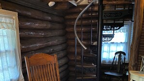 Ein Blockhaushaus-Interieur mit einem hölzernen Schaukelstuhl, einem kleinen Tisch mit einer gehäkelten Tischdecke, einem weißen, flauschigen Teppich und einer Wendeltreppe.