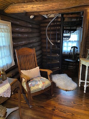 A log cabin interior with a wooden rocking chair, a small table with a crocheted tablecloth, a white furry rug, and a spiral staircase.