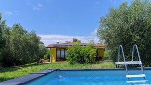 Une piscine avec un escalier, entourée de verdure et bordée d’un bâtiment jaune au toit recouvert de tuiles.