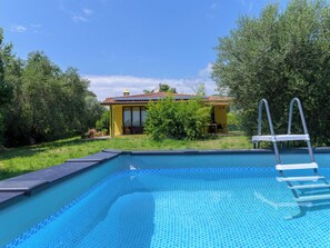 Ein Schwimmbecken mit Leiter, umgeben von Grünpflanzen und einem gelben Gebäude mit einem Kachelziegeldach.