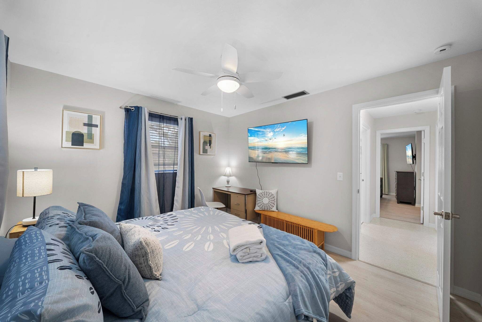 Un dormitorio con cama, escritorio, silla, lámpara de pie, ventilador de techo y puerta que da a otra habitación.