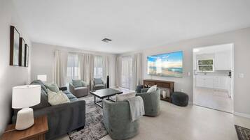 A modern living room with a sofa set, a coffee table, and a TV. There is a kitchen area visible through an open door.