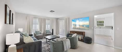 A modern living room with a sofa set, a coffee table, and a TV. There is a kitchen area visible through an open door.