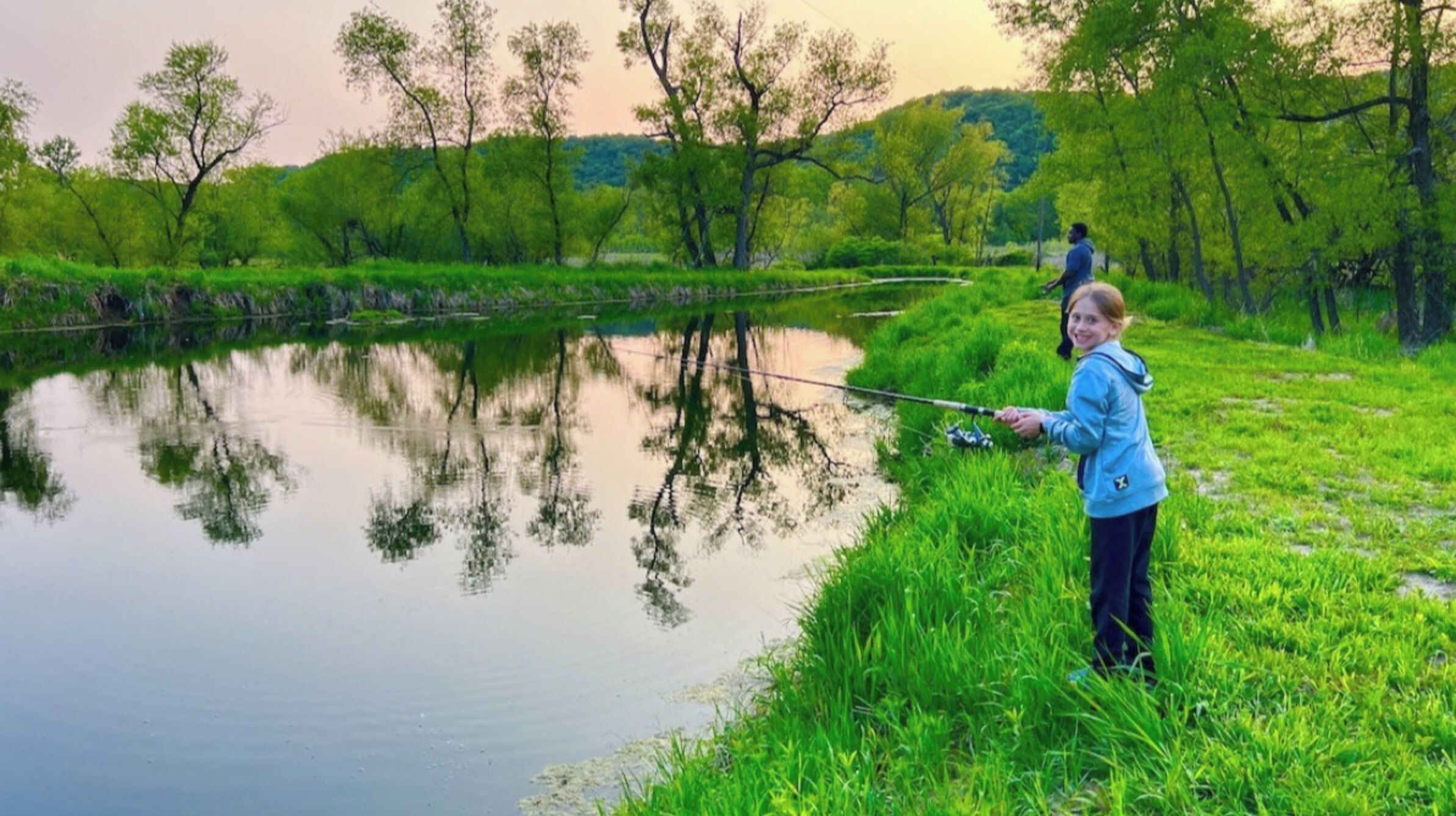 Una persona pescando en un río tranquilo, con abundante vegetación y árboles al fondo.