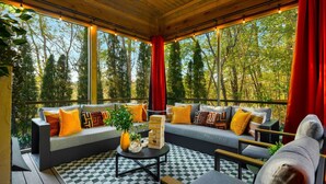 A covered outdoor seating area with a checkered floor, a sofa with cushions, a small table with a vase and blocks, and a view of trees through large windows. - Sleeps 20 • 15 Mins to DT AVL • Sauna • Hot Tub! (Fletcher)