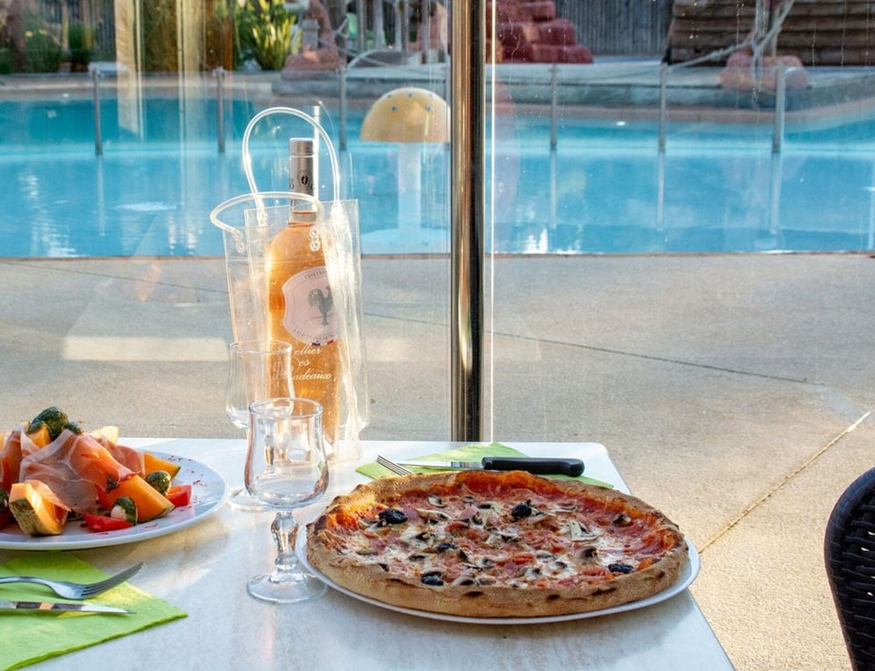 A dining table with a pizza, a bottle of wine, and a plate of vegetables by a poolside.