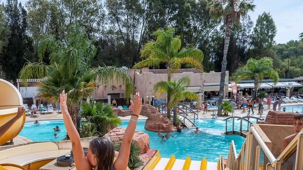 Area piscina con diverse scivoli d'acqua, palme e persone che nuotano.