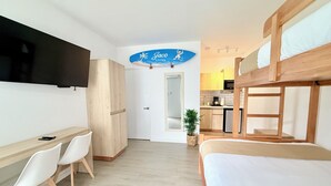 Une pièce comprenant un lit superposé, un bureau avec des chaises, une télévision, un coin cuisine et une porte donnant sur une autre pièce.