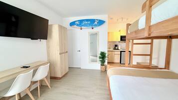 A room with a bunk bed, a desk with chairs, a television, a kitchen area, and a door leading to another room.