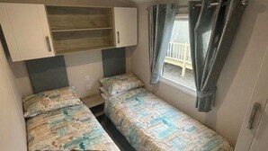 A small, compact room with two single beds, a window with curtains, and a kitchenette area. - Golden Palm Resort - Palm Skye PG155 (Chapel Saint Leonards)