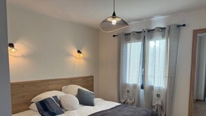 Un dormitorio con cabecera de madera, cama con ropa de cama blanca y gris, dos luces fijadas a la pared y una ventana con cortinas grises.