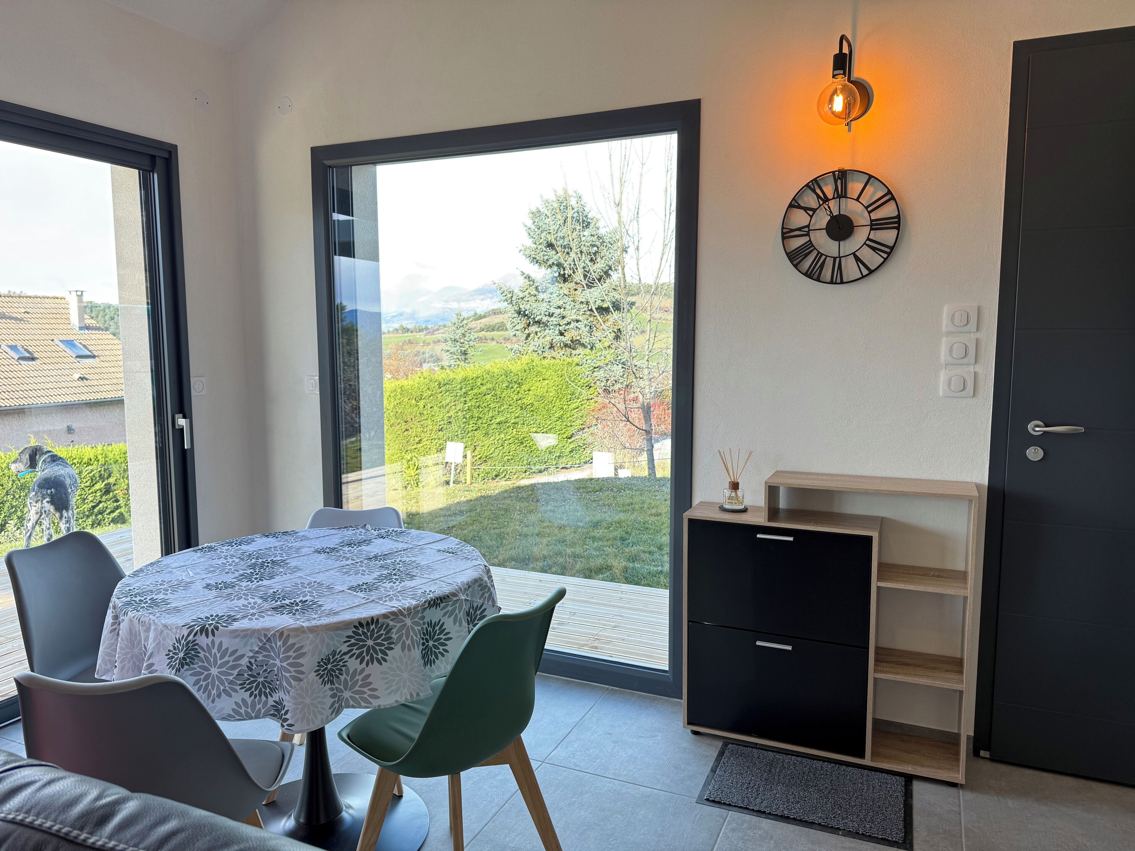 A room with a table, chairs, a clock, a door, and a view of the outdoors.