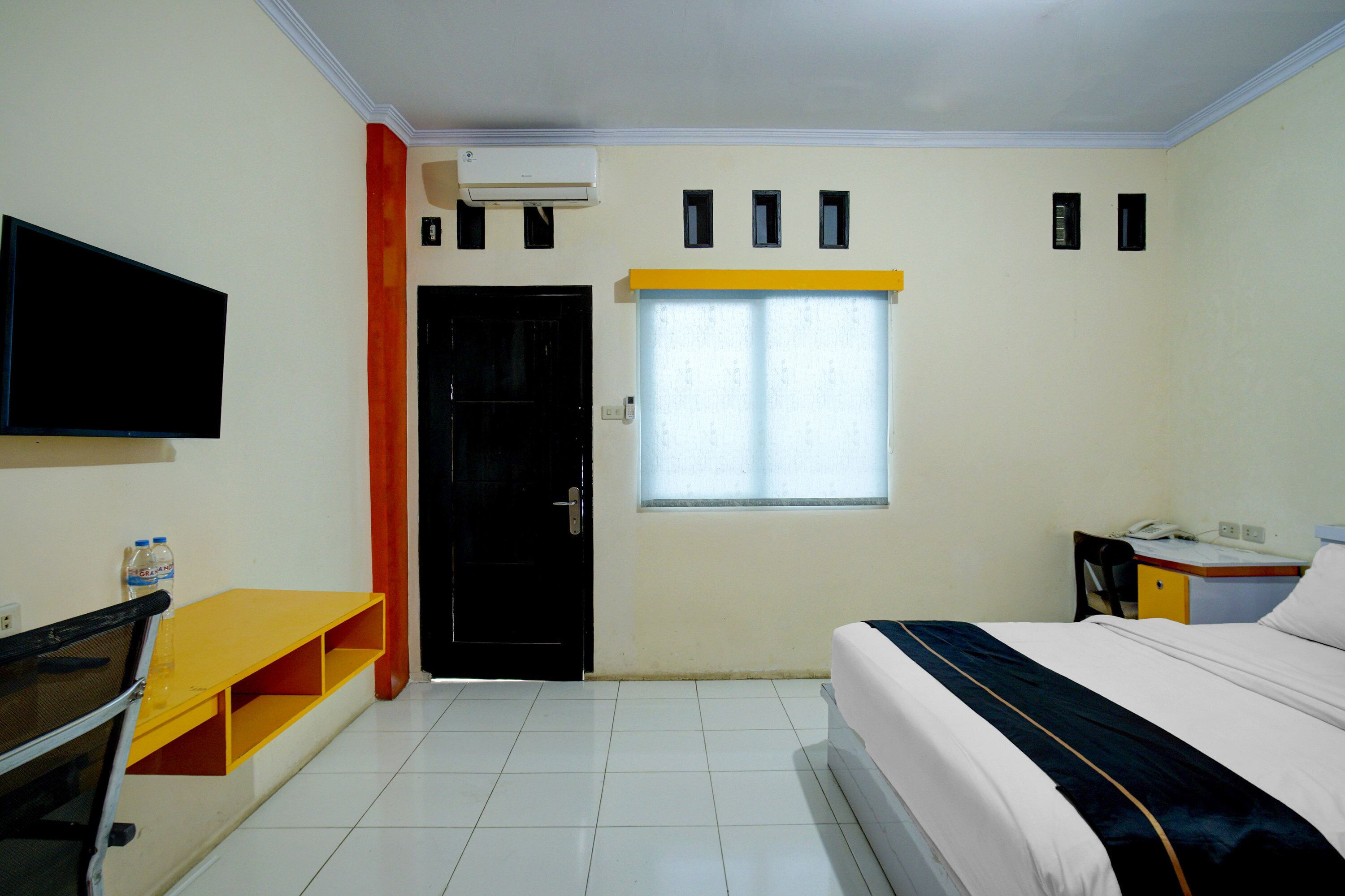 Habitación de hotel con cama, escritorio, silla, televisor, ventana con persianas y aire acondicionado.