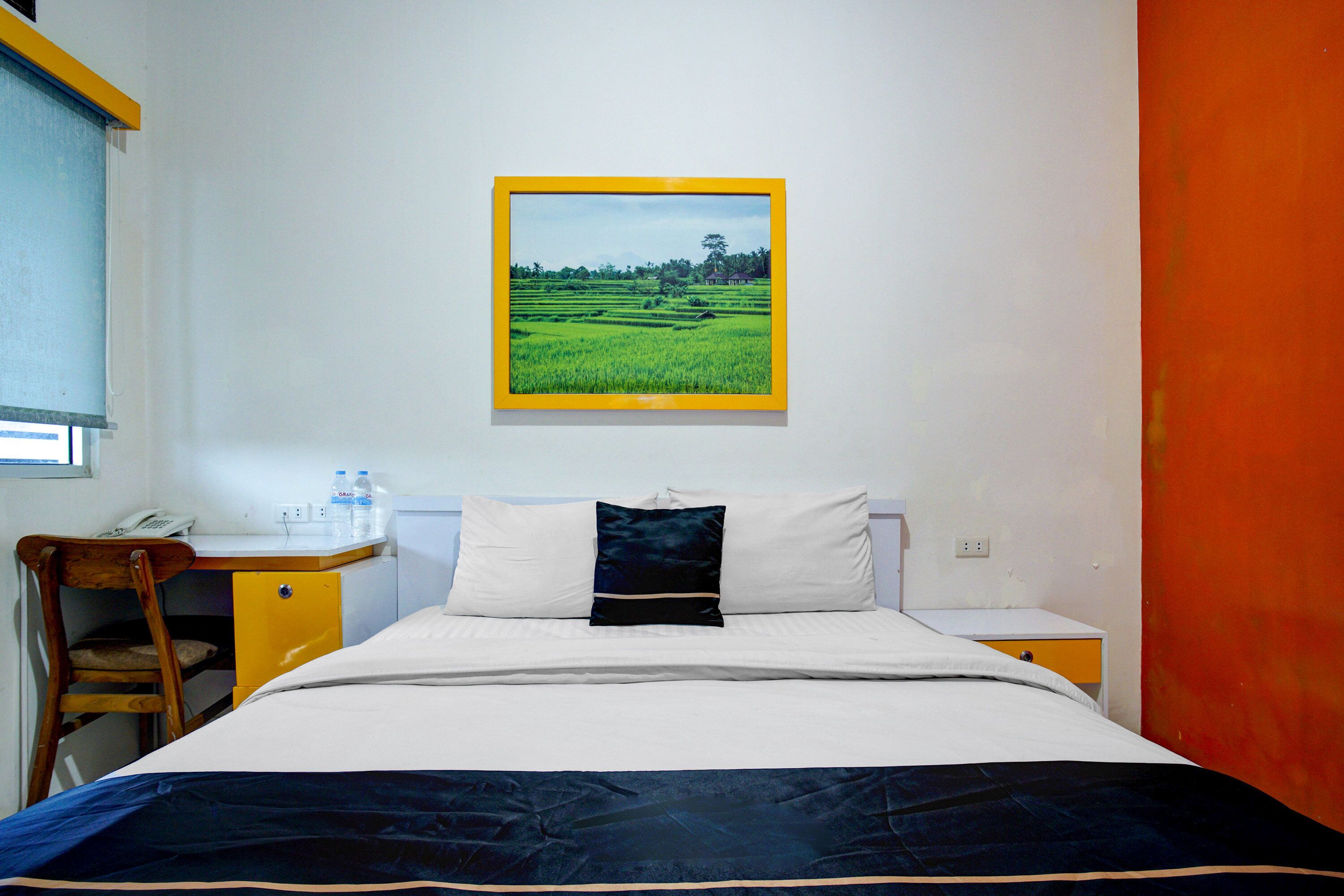 Ein Hotelzimmer mit einem Bett, einem Schreibtisch mit Telefon, einem kleinen Tisch mit Wasserflaschen und einem Bild einer grünen Wiese an der Wand.
