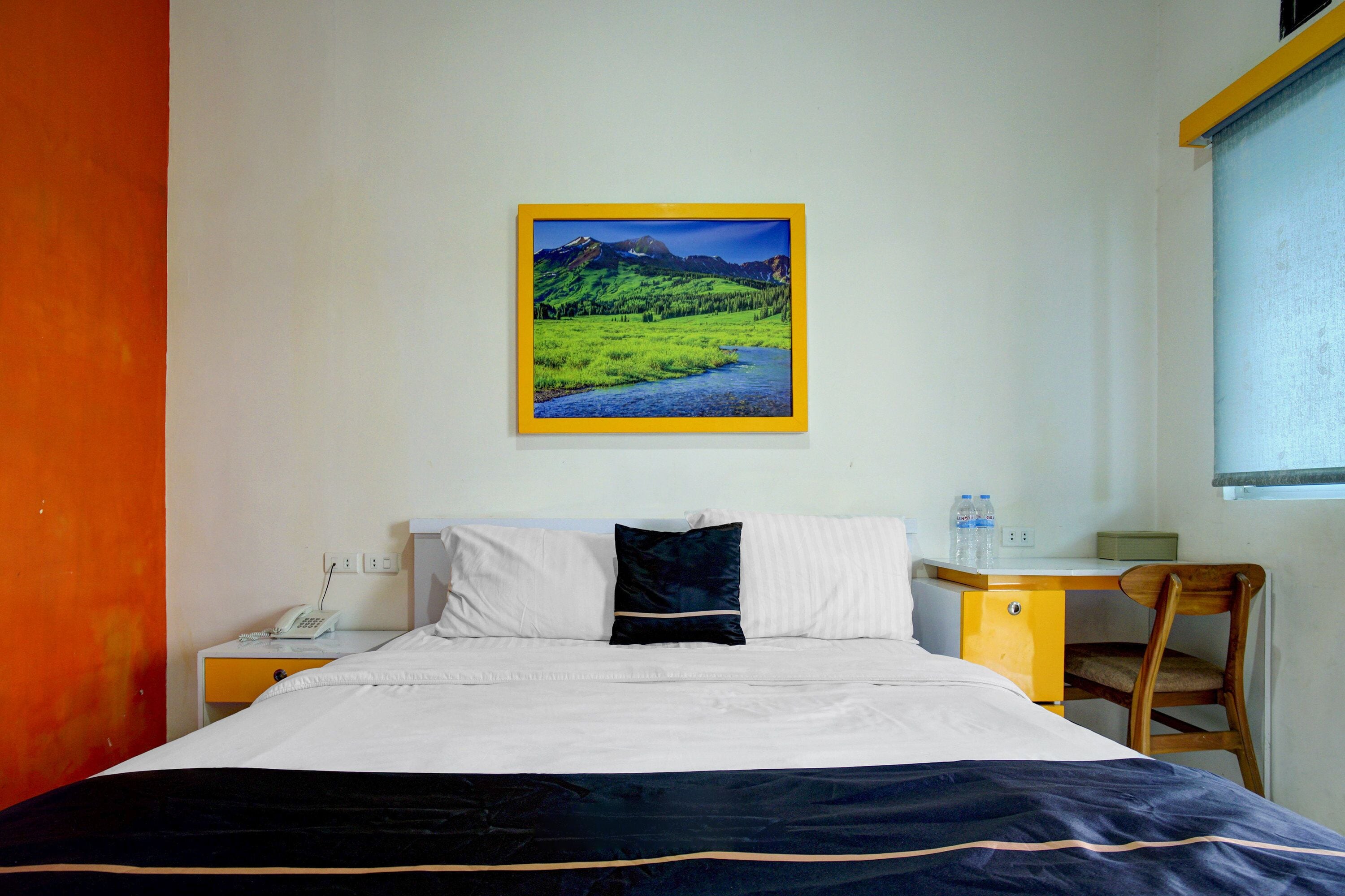 A hotel room with a bed, a nightstand with a phone, a desk with a chair, and a framed landscape painting on the wall.