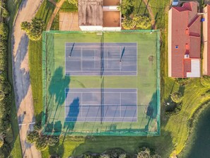 Luftaufnahme eines Tennisplatzes, umgeben von Grünflächen und einem Gebäude mit rotem Dach.