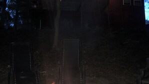 Een kampvuur in een metalen vuurkorf, met een huis op de achtergrond.