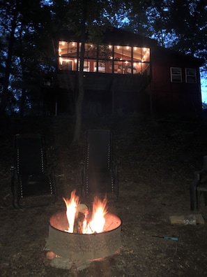 Een kampvuur in een metalen vuurkorf, met een huis op de achtergrond.
