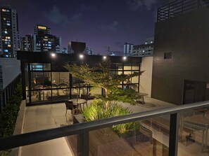 A rooftop terrace with outdoor seating, a glass wall, and a view of the city skyline at dusk. - Flat Sol & Praia Move Tambaú (João Pessoa)
