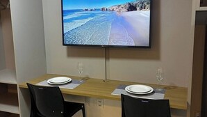 A dining area with a TV mounted on the wall displaying a beach scene, two chairs, and a table set for two. - Flat Sol & Praia Move Tambaú (João Pessoa)